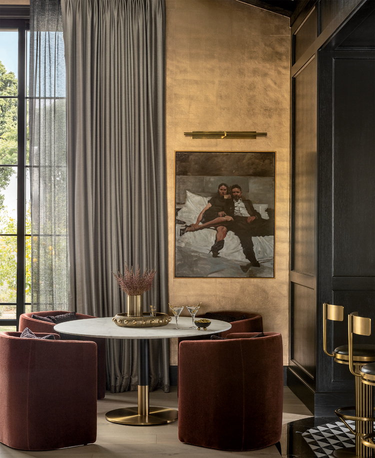 A round table with four burgundy chairs sits by tall gray curtains, a gold accent wall, and a framed painting of a couple above the table. Gold decor and barstools are visible.