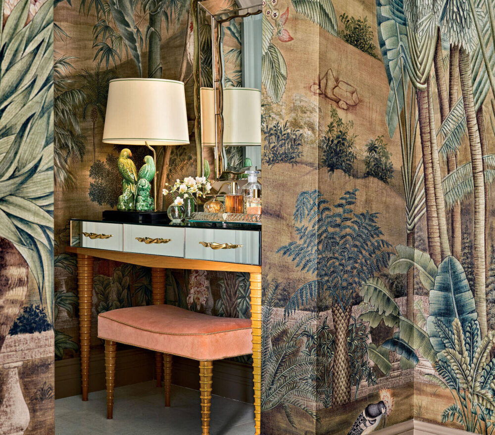 A vanity with a light blue drawer, gold accents, a lamp, drinks, and a pink bench sits in a room with tropical-themed wallpaper and a mirror.