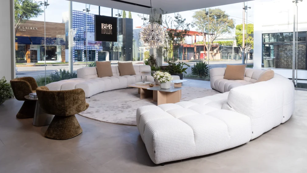 Modern showroom with large curved white sofas, two brown chairs, coffee tables, and a round rug, surrounded by floor-to-ceiling glass windows with a view of the street outside.