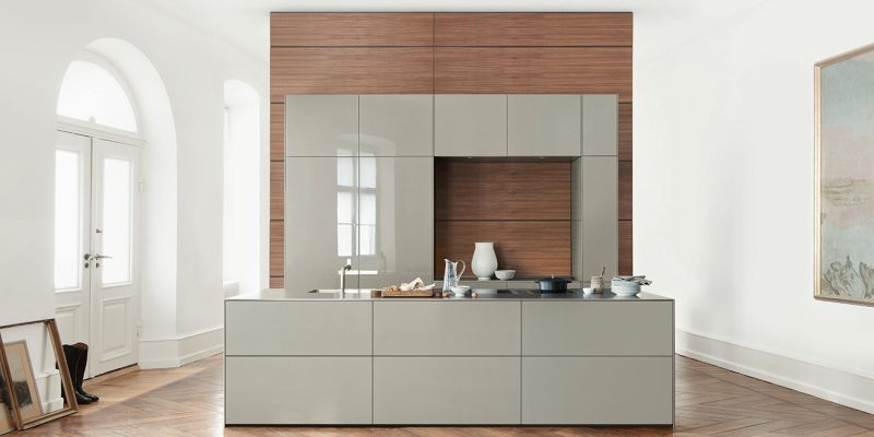 Modern kitchen with glossy gray cabinets, a central island, wood panel accents, and minimal decor in a bright room with large windows and herringbone wood flooring.