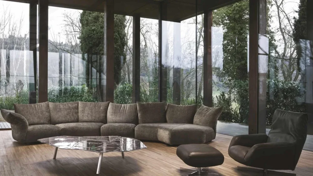 Modern living room with a large brown sectional sofa, angular coffee table, and dark armchair with ottoman, set against floor-to-ceiling windows overlooking trees.