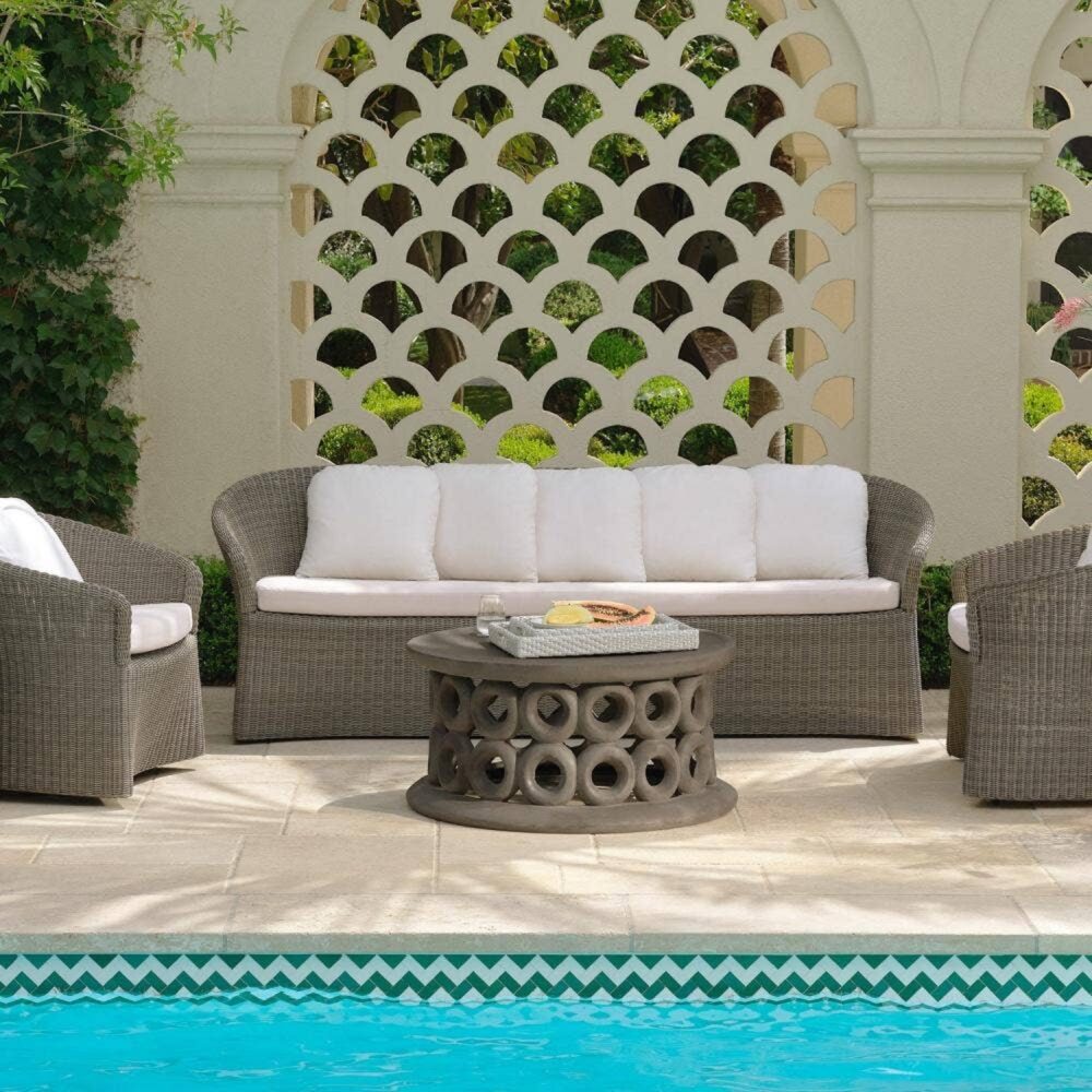 Wicker outdoor furniture with white cushions surrounds a round table with snacks, set on a stone patio by a swimming pool. Patterned wall and greenery in the background.