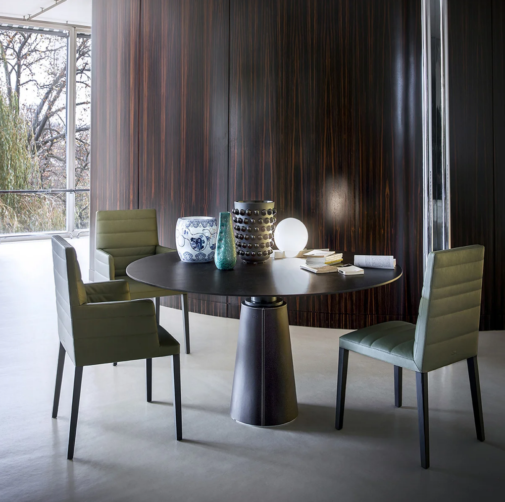 Round black table with three green chairs, various decorative objects, books, and a lamp on top; dark wood paneled wall and large window in the background.