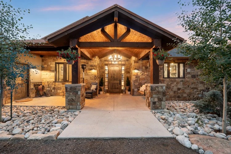 A stone and wood house with a covered entrance, hanging plants, pathway, and surrounding landscaping with rocks and small trees at dusk.