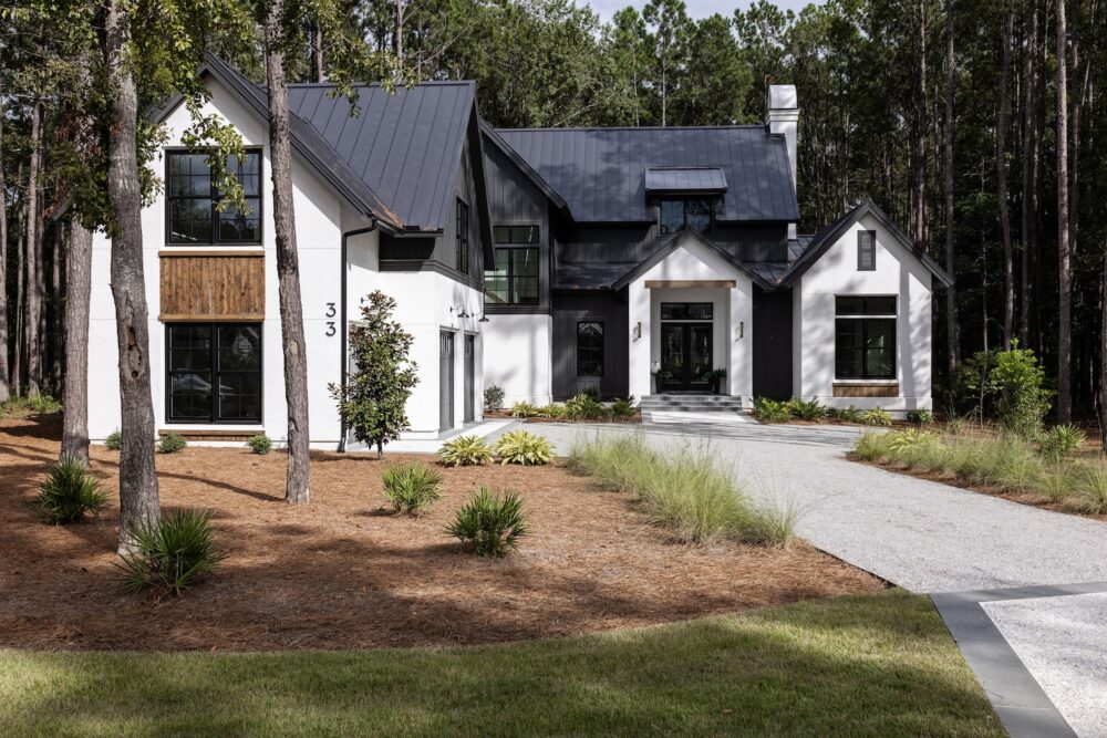 Modern two-story house with black metal roof, white and gray exterior walls, large windows, and a gravel driveway surrounded by pine trees and landscaped yard.