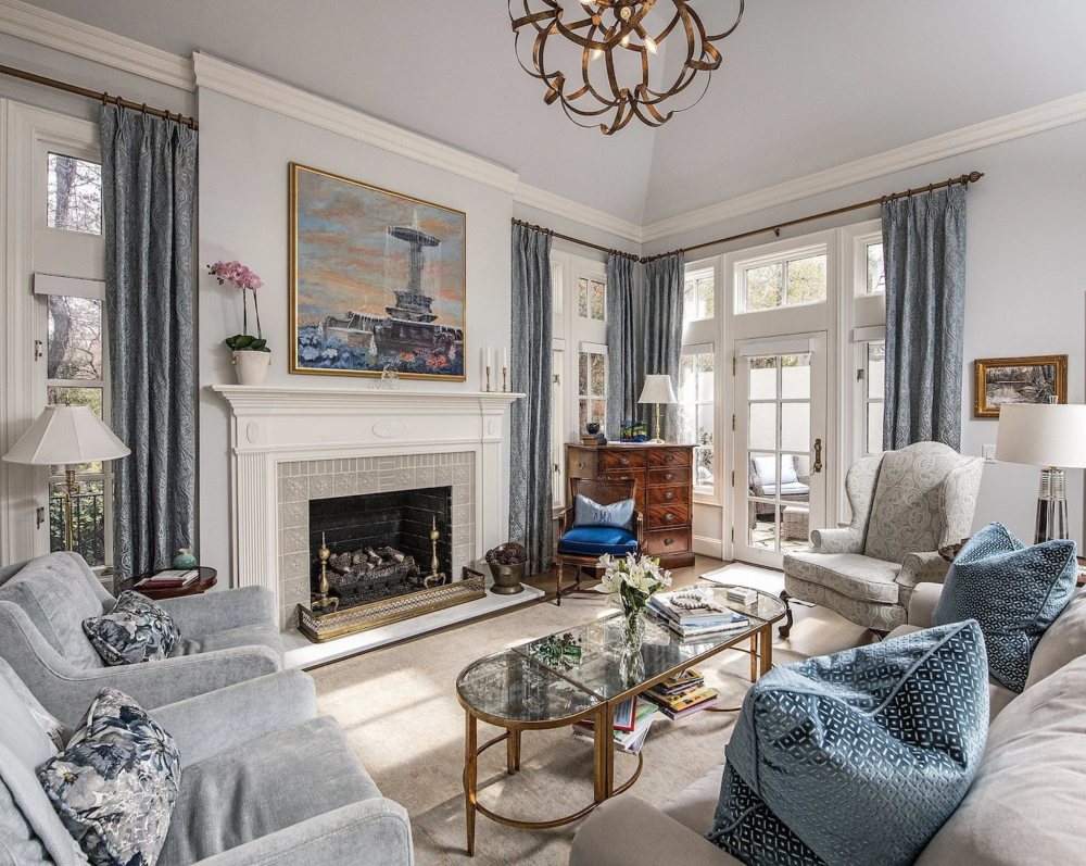 Elegant living room with a fireplace, blue-grey sofas, glass coffee table, armchairs, large windows with drapes, and a chandelier. Artwork hangs above the mantel.