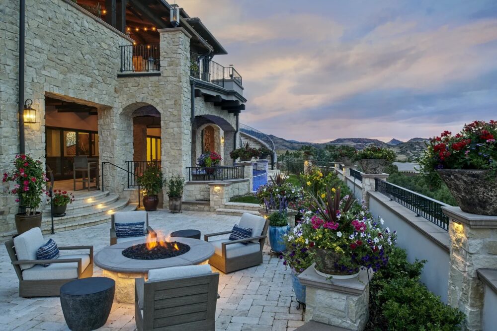 Stone patio with cushioned chairs around a firepit, colorful flowers in planters, and a large house with balconies, set against a backdrop of rolling hills at sunset.