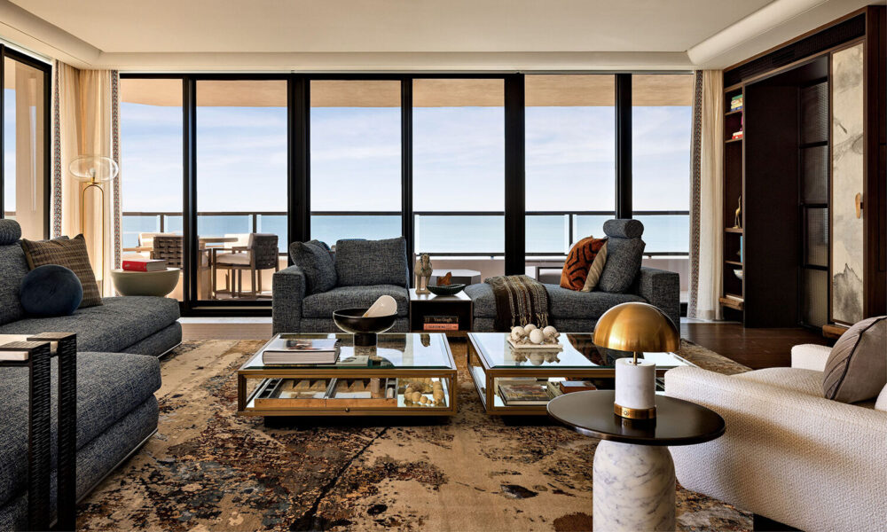 Modern living room with blue sofas, glass coffee tables, and floor-to-ceiling windows overlooking a balcony with a sea view. Shelves and decor items are arranged throughout the space.
