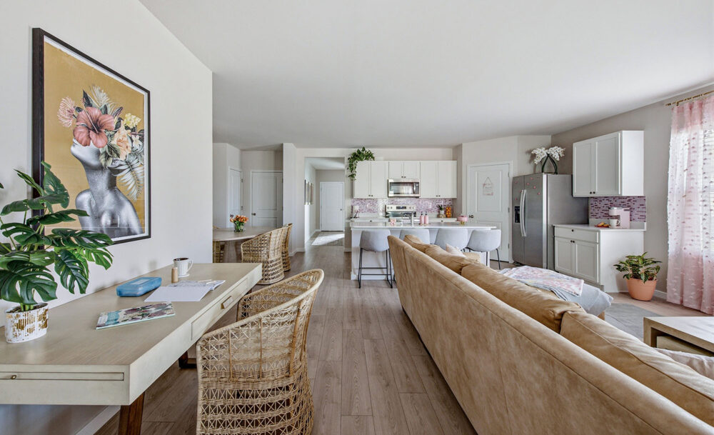 Open-plan living area with a desk, wicker chairs, beige sofa, plants, wall art, and a modern kitchen with white cabinets and stainless steel appliances.