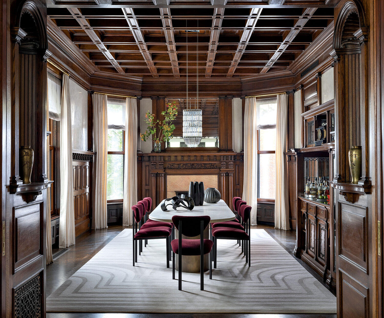 Formal dining room with dark wood paneling, a large rectangular table surrounded by burgundy chairs, modern chandelier, fireplace, large windows, and decorative vases on built-in shelves.