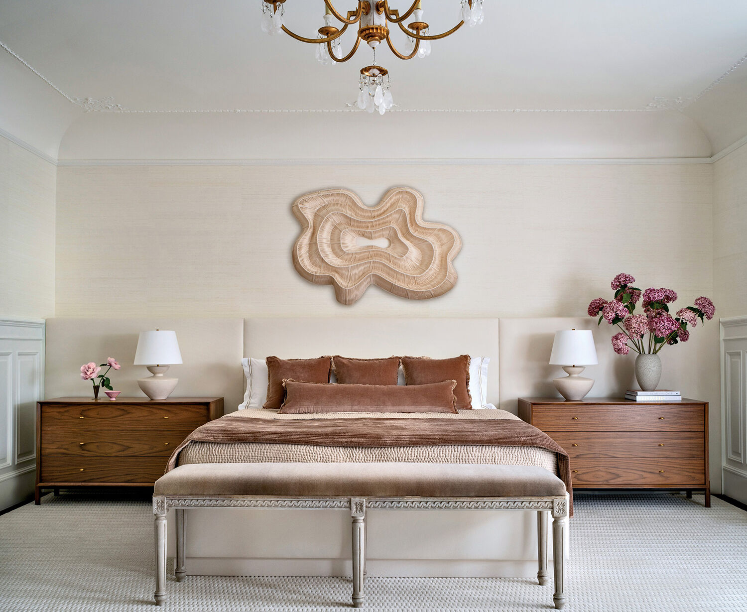 A neatly made bed with brown bedding sits between two wooden nightstands, each with a lamp and flowers. Abstract wall art and a chandelier decorate the neutral-toned bedroom.