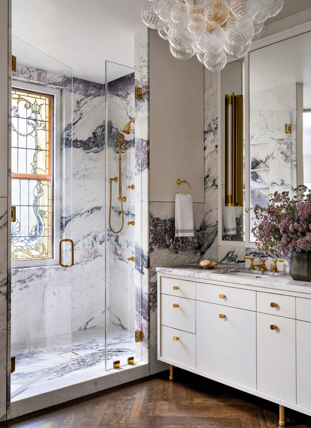Elegant bathroom with marble walls, glass shower, gold fixtures, white vanity, and a decorative ceiling light. A vase of purple flowers sits on the counter near a stained glass window.