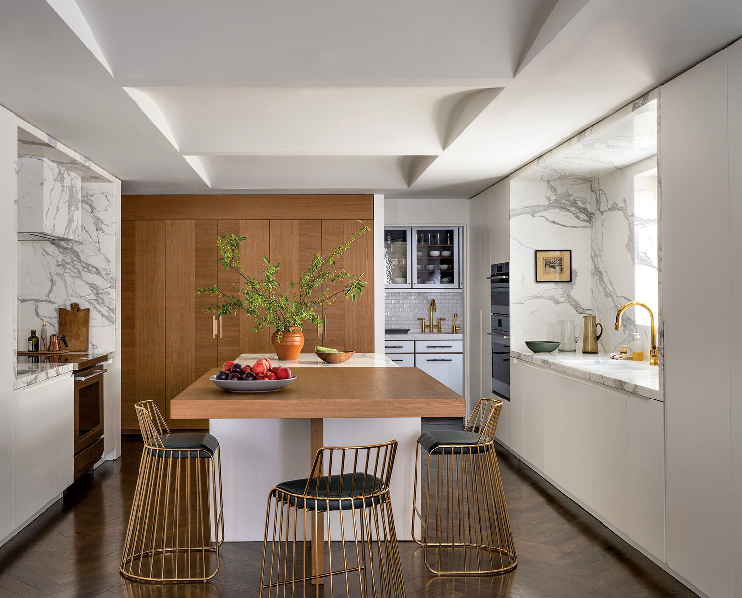 Modern kitchen with wood and white cabinetry, marble surfaces, a central island with bar stools, gold accents, and a bowl of fruit and vase with greenery on the island.