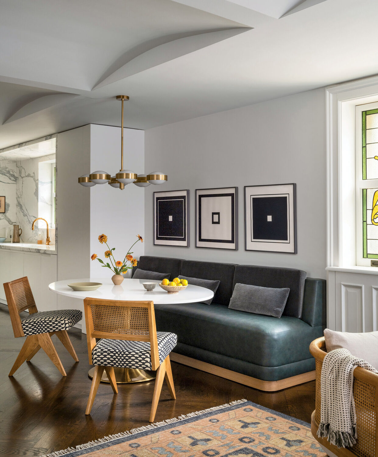 Modern dining nook with a round white table, three chairs, dark green bench, wall art, pendant light, and kitchen visible in the background. Flowers and fruit are on the table.