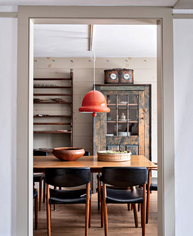 View through a doorway into a dining room with a wooden table, black chairs, orange pendant light, shelves, and a rustic cabinet with clocks on top.