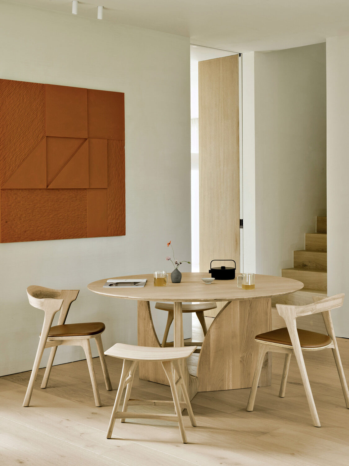 A round wooden dining table with three chairs, a small teapot, glasses, and a flower vase in a minimal room with a geometric wall art and light wood flooring.