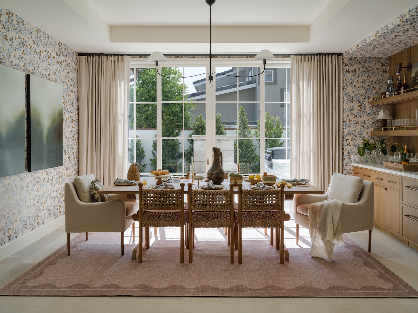 A dining room with a wooden table, eight woven chairs, beige armchairs, a pink rug, large window, patterned wallpaper, and built-in shelves set for a meal.