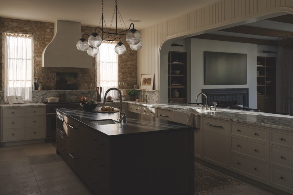 Spacious kitchen with marble countertops, large island, modern pendant lights, stainless appliances, and a view into a living area with a TV and fireplace. Natural light enters through tall windows.