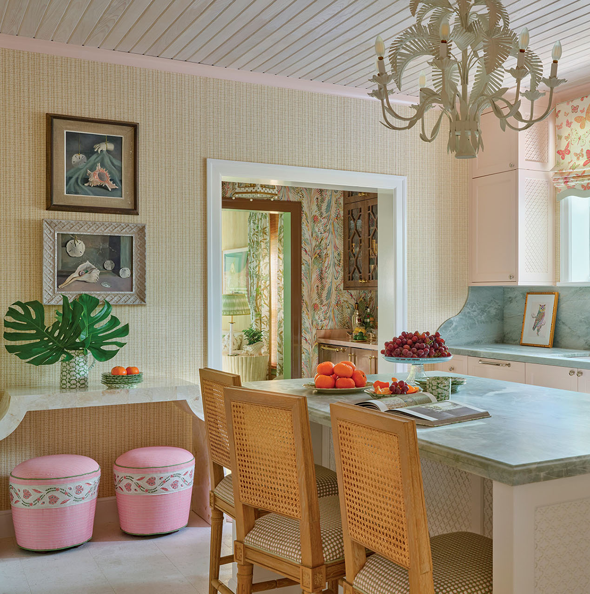Colorful kitchen with rattan chairs, marble island, pink accent stools, tropical leaf decor, fruit on counter, and framed art on patterned wallpaper walls. Doorway leads to another decorated room.