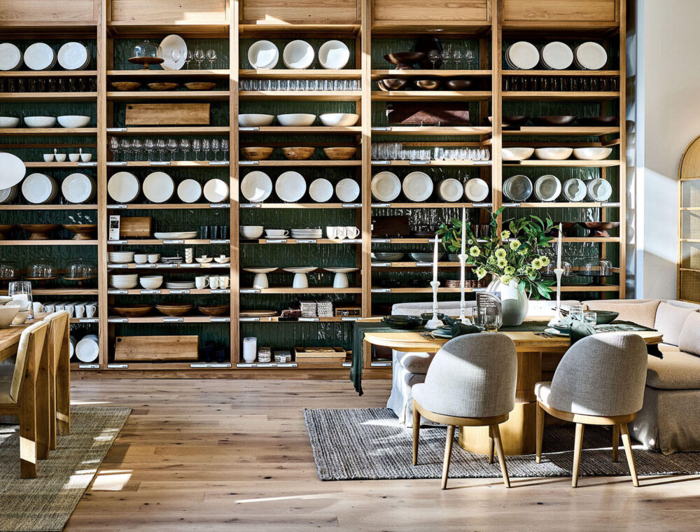 Open wooden shelves display neatly arranged dishes, glassware, and bowls. Modern dining and living area with natural light, gray chairs, wood tables, and a vase of flowers on a rug.