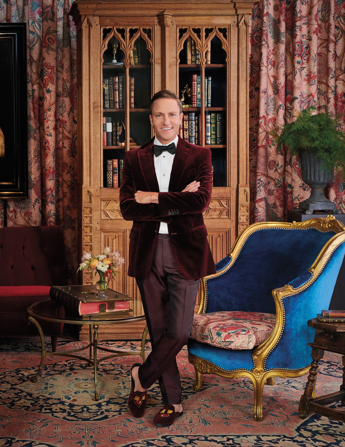 A man in a velvet tuxedo stands with arms crossed in an ornate, library-like room with patterned curtains, antique furniture, and bookshelves.