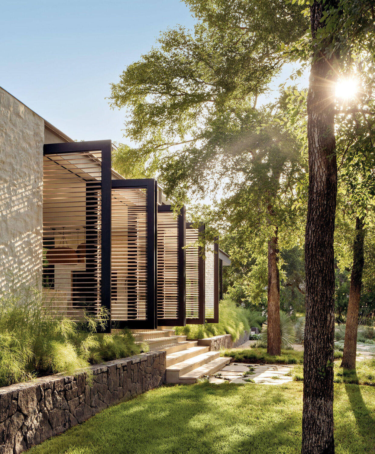 Modern house exterior with horizontal wooden slats, stone retaining wall, steps, and lush green landscaping, with sunlight streaming through trees.
