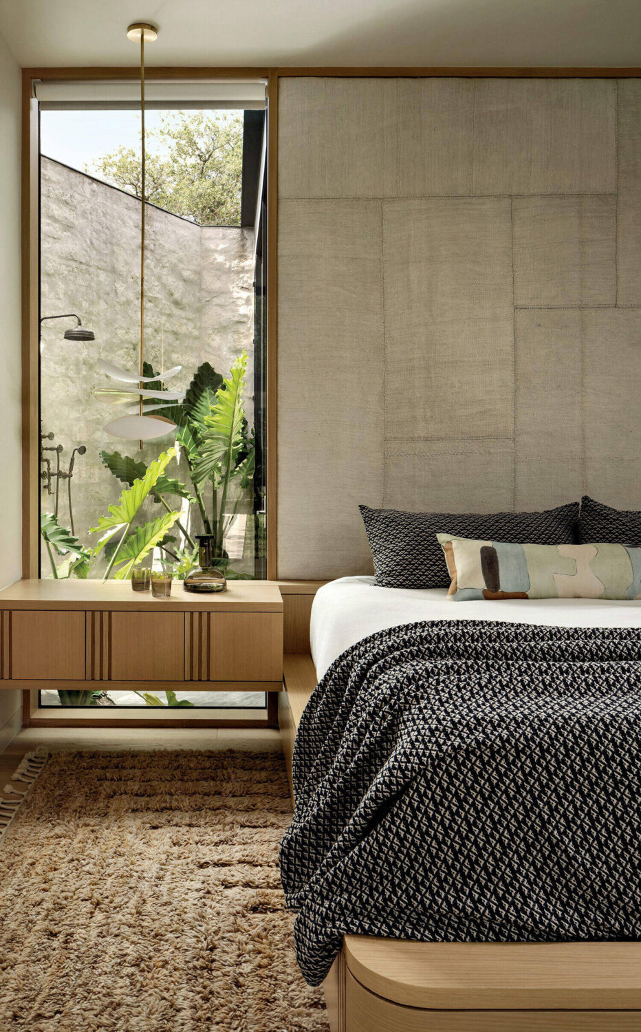 Modern bedroom with a wooden bed, black patterned bedding, a pendant light, a bedside table, and a large window showing greenery and an outdoor shower.