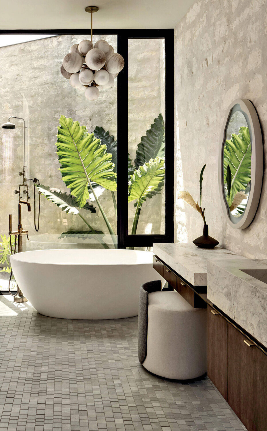 Modern bathroom with a white freestanding tub, round mirror, marble counter, and large window showing green tropical plants outside.