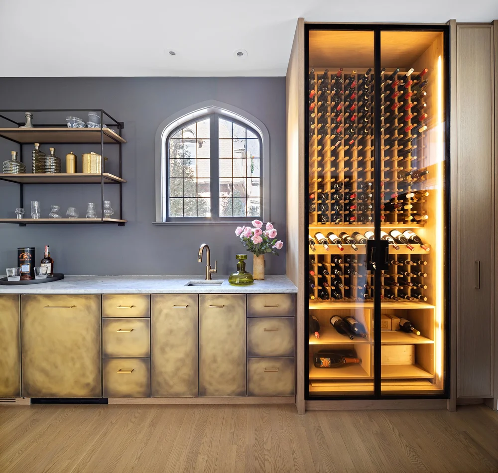 Modern home bar with brass cabinets, open shelves, a marble countertop, a gold faucet, a window, and a tall, illuminated wine fridge filled with bottles. Pink flowers in a vase are on the counter.