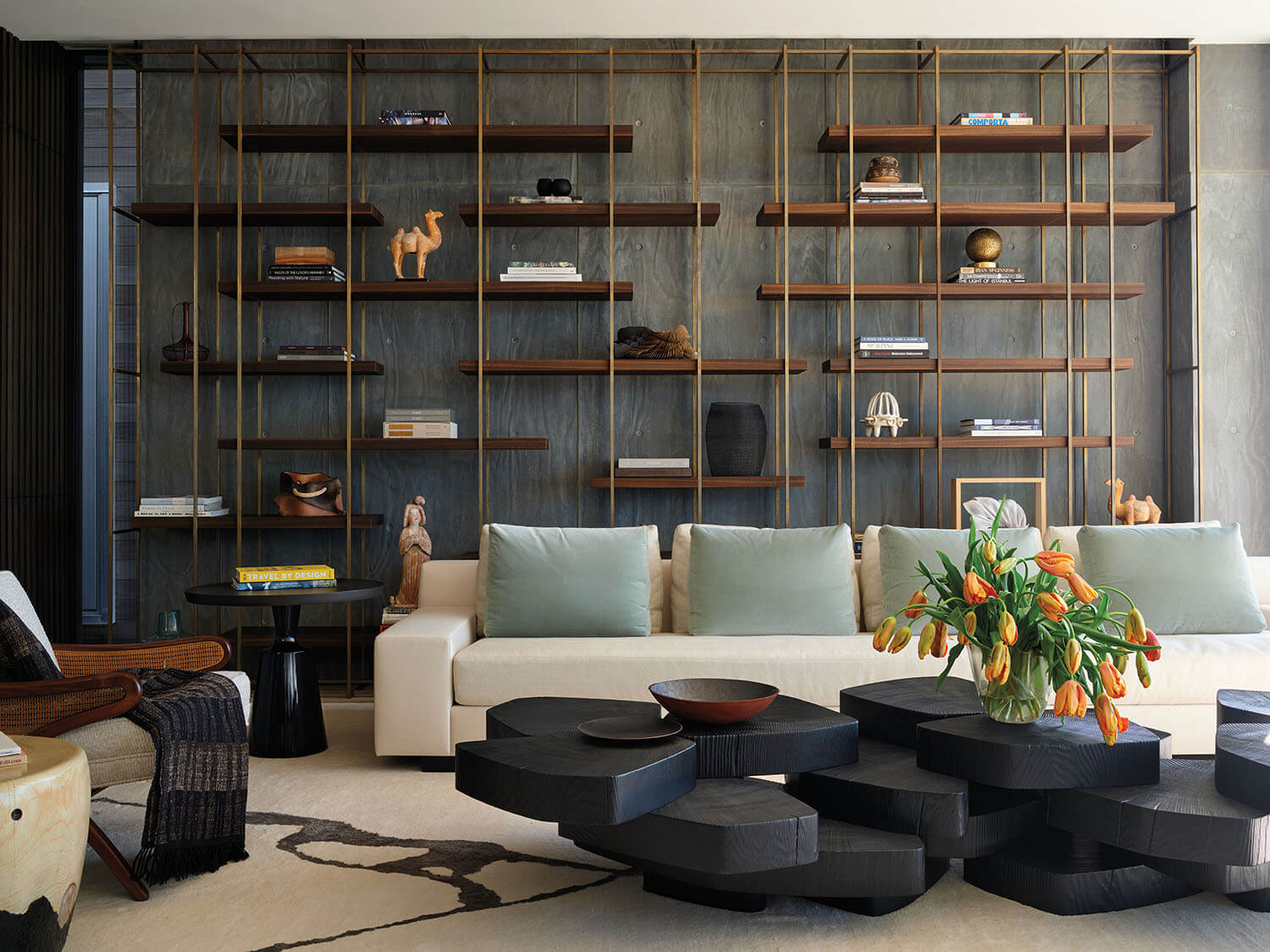 Modern living room with a white sofa, green cushions, black tiered coffee tables, orange tulips in a vase, and a large wall-mounted bookshelf with various decorative items.