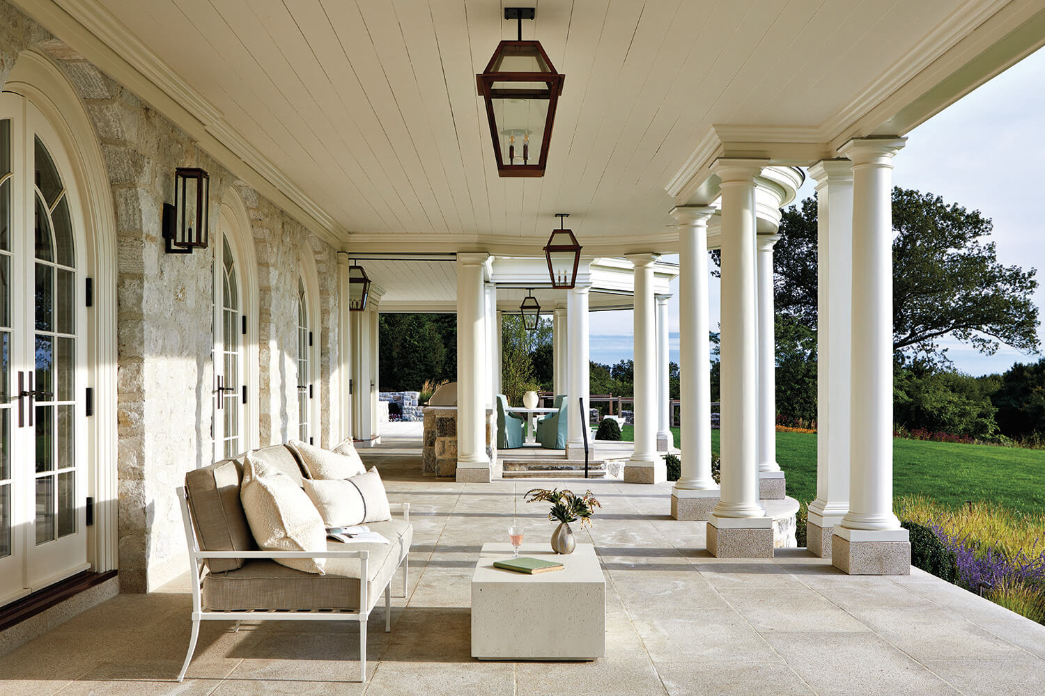 Spacious covered patio with stone flooring, white columns, outdoor seating, and a table, overlooking a green lawn and trees under a bright sky.