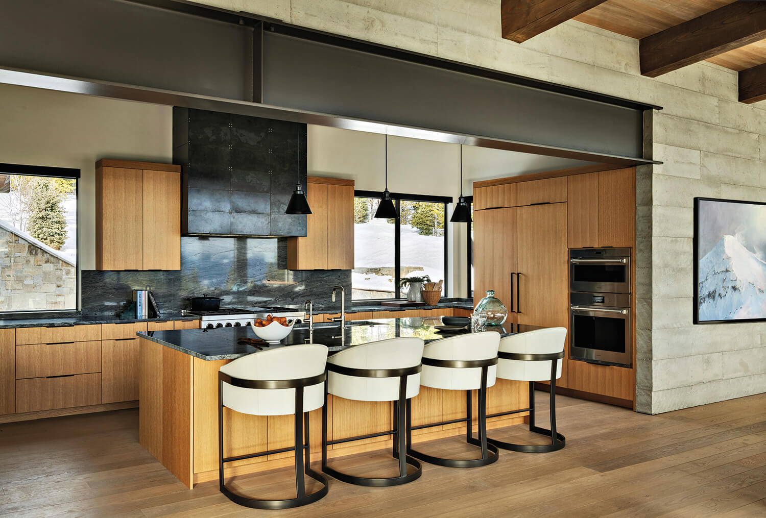 Modern kitchen with wood cabinets, a black countertop island, four white barstools, built-in appliances, and large windows with a snowy outdoor view.