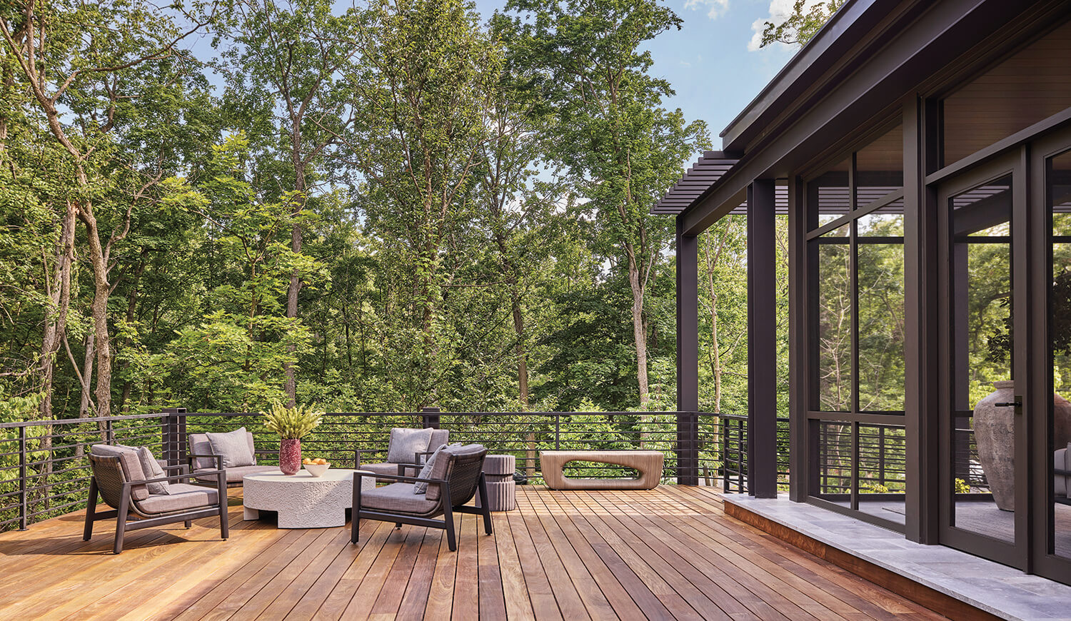 Spacious wooden deck with black railings, modern outdoor furniture, and a view of tall green trees surrounding the area on a sunny day.