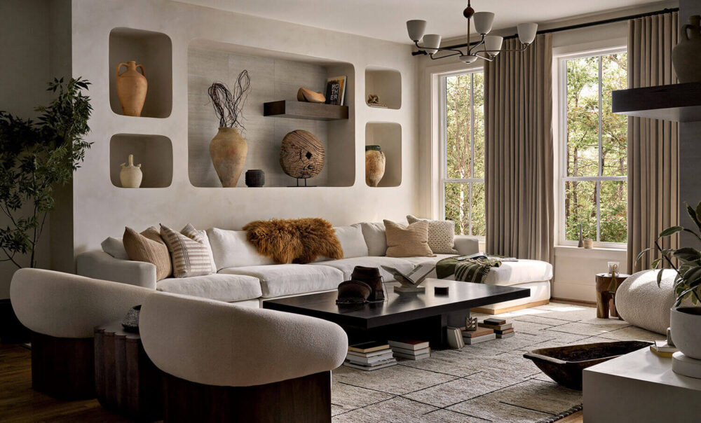 Modern living room with a large white sectional sofa, round chairs, wall alcoves displaying pottery, a black coffee table, books, and large windows with beige curtains letting in natural light.