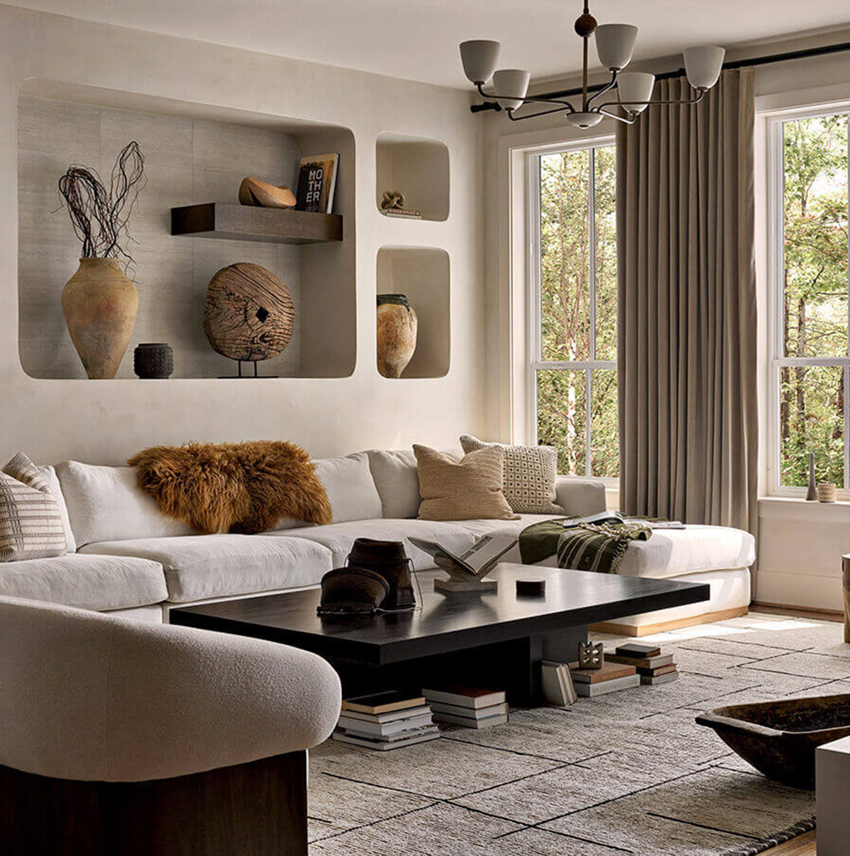 Modern living room with a white sectional sofa, black coffee table, neutral decor, built-in wall shelves displaying pottery, and large windows with beige curtains letting in natural light.