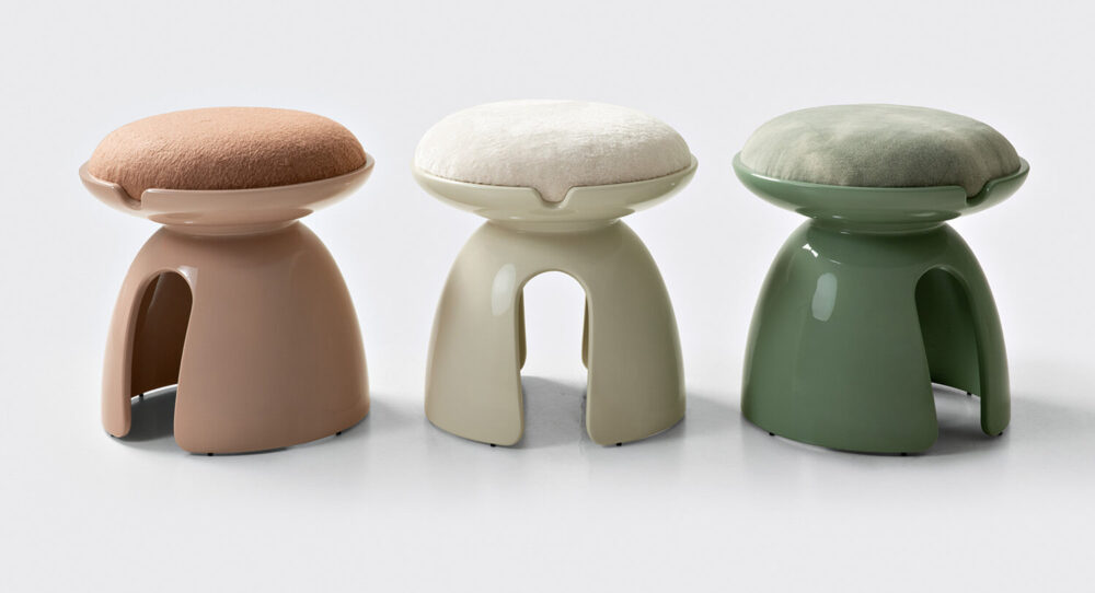 Three modern stools with glossy, curved bases in beige, off-white, and green, each topped with a round, cushioned seat in coordinating pastel colors, are placed side by side on a white background.