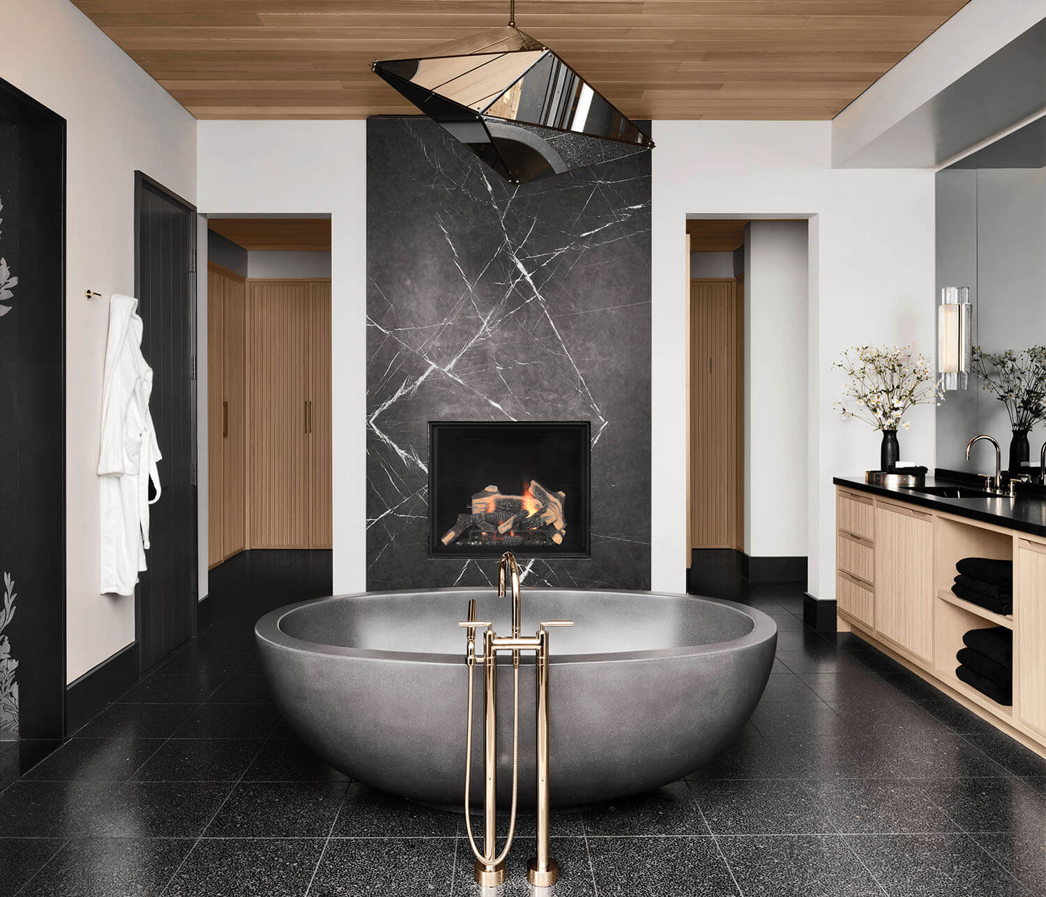 Modern bathroom with a freestanding oval bathtub, black marble fireplace, black tile floor, wooden ceiling, and light wood cabinetry.