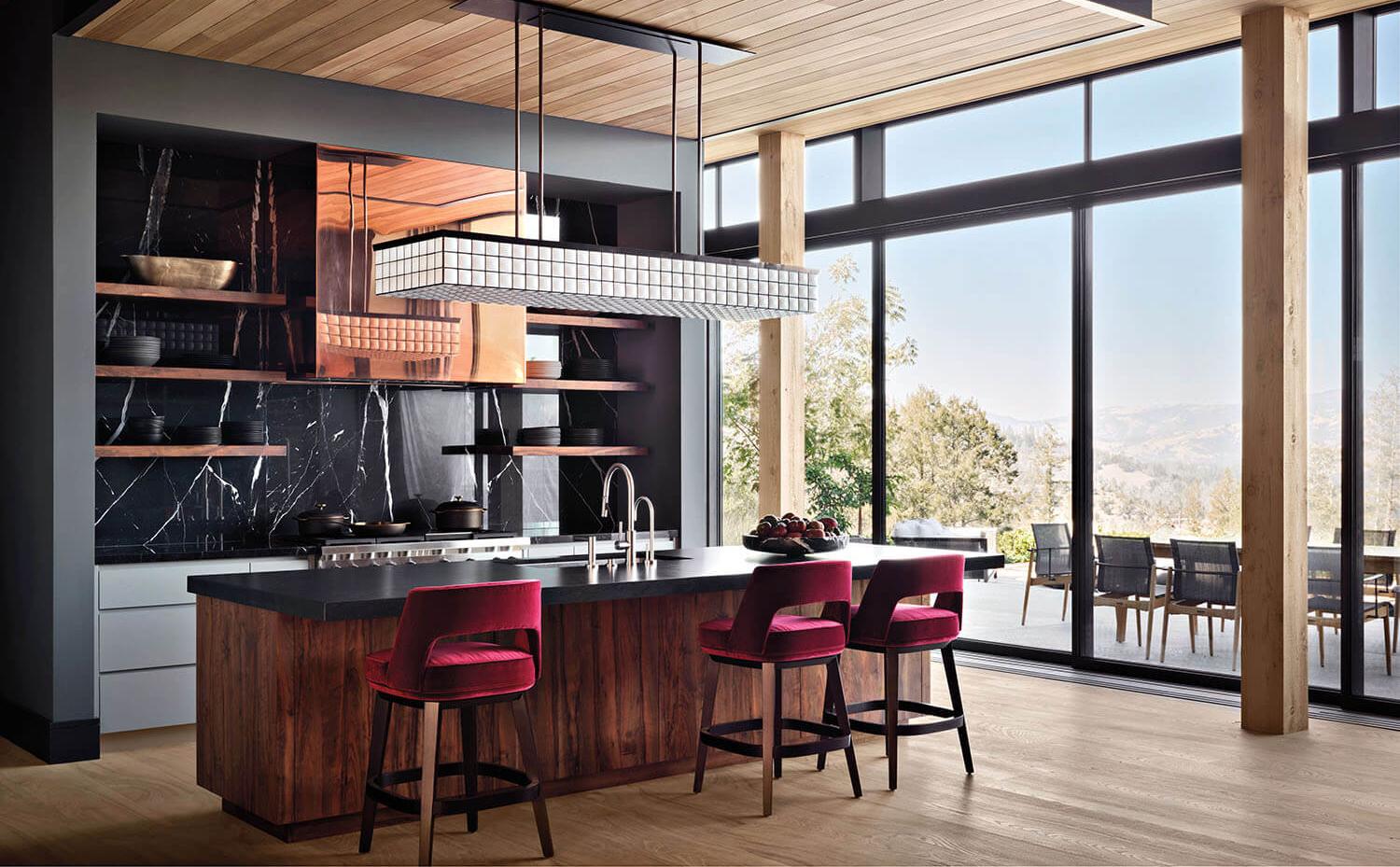 Modern kitchen with a wood island, three red barstools, black marble backsplash, open shelves, pendant light, and large windows overlooking an outdoor dining area and scenic landscape.
