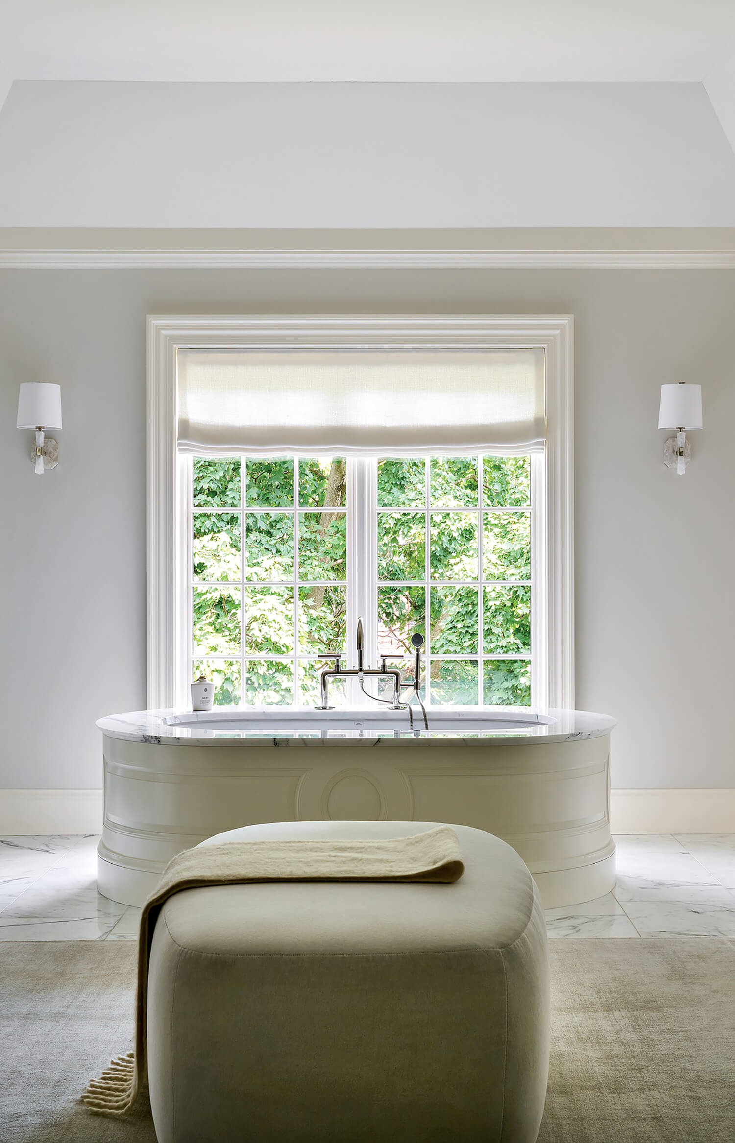 A white freestanding bathtub sits under a large window with a leafy view, flanked by two wall sconces, with an ottoman in the foreground.