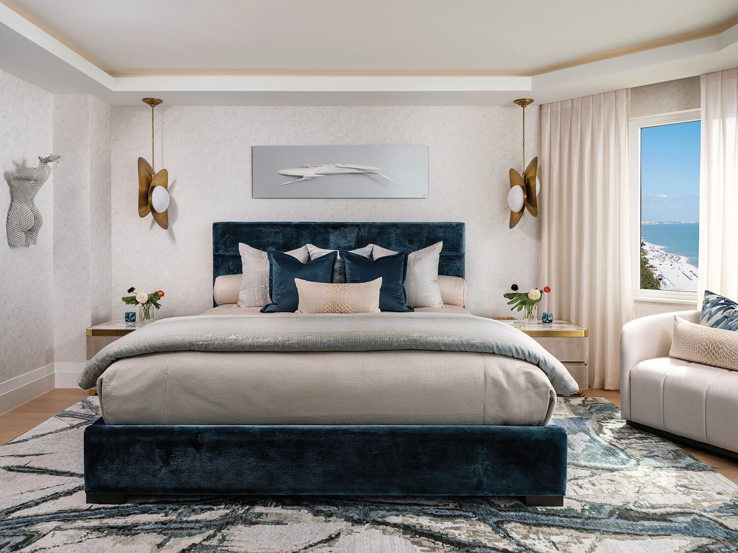 Modern bedroom with a blue velvet bed, beige and blue bedding, gold light fixtures, nightstands with flowers, abstract wall art, and a window showing an ocean view.