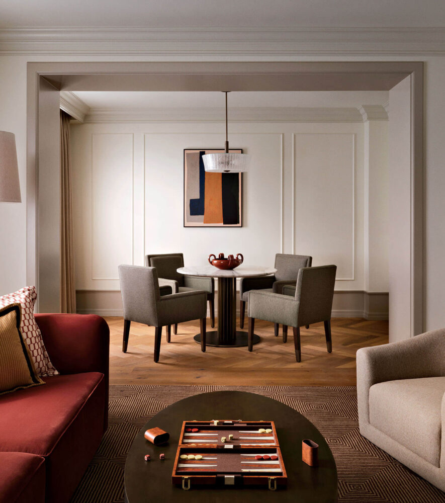 A modern living room with a maroon sofa, a round coffee table holding a backgammon board, and a dining area with four gray chairs around a circular table.
