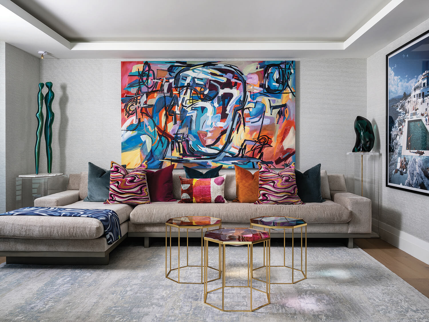 Modern living room with a large abstract face painting, beige sectional sofa with colorful pillows, geometric glass coffee tables, and contemporary sculptures on display.