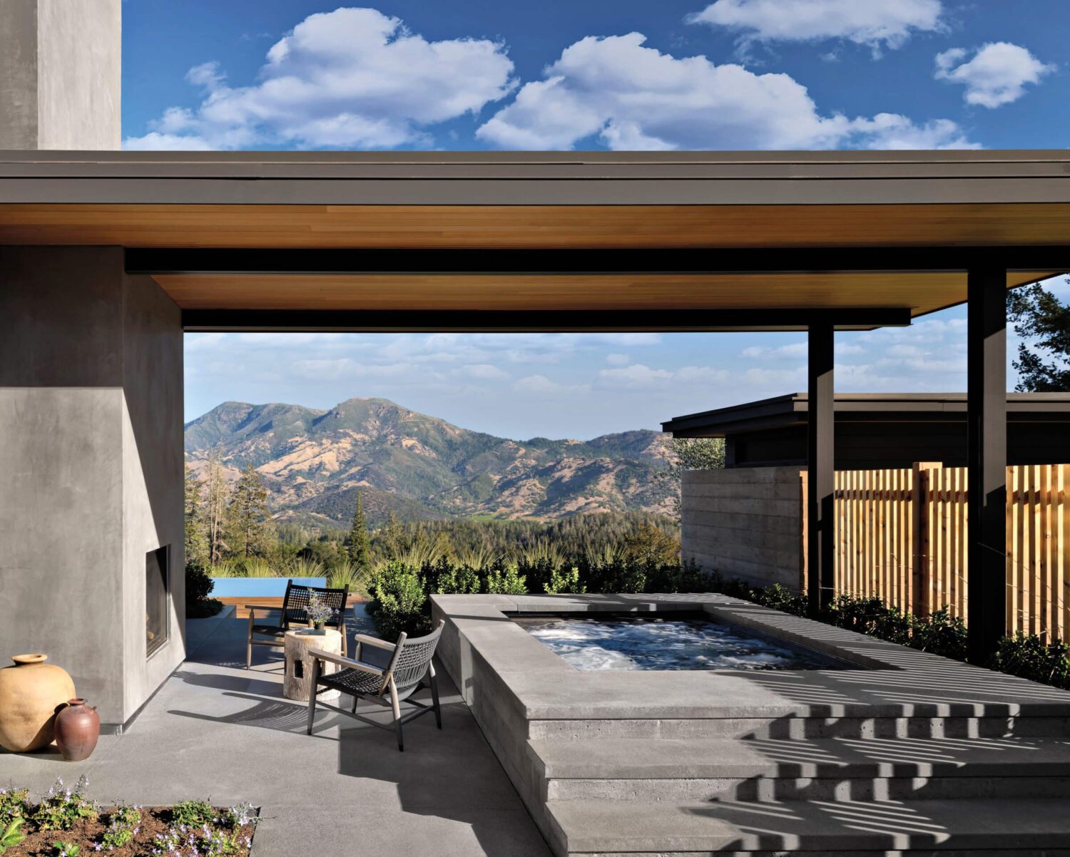 Modern patio with a hot tub, chairs, and a table, partially covered by a roof, overlooking distant mountains under a blue sky with clouds.