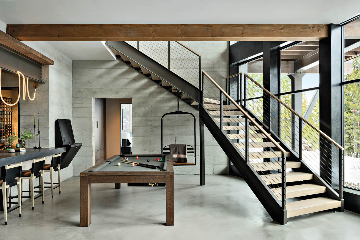 Modern interior with a wooden staircase, pool table, bar stools, and large windows allowing natural light into the space.