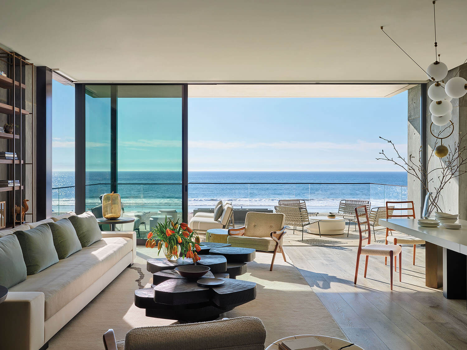Modern living room with large windows overlooking the ocean, featuring contemporary furniture, a white sofa, and wooden dining table. Bright and airy atmosphere with natural sunlight.