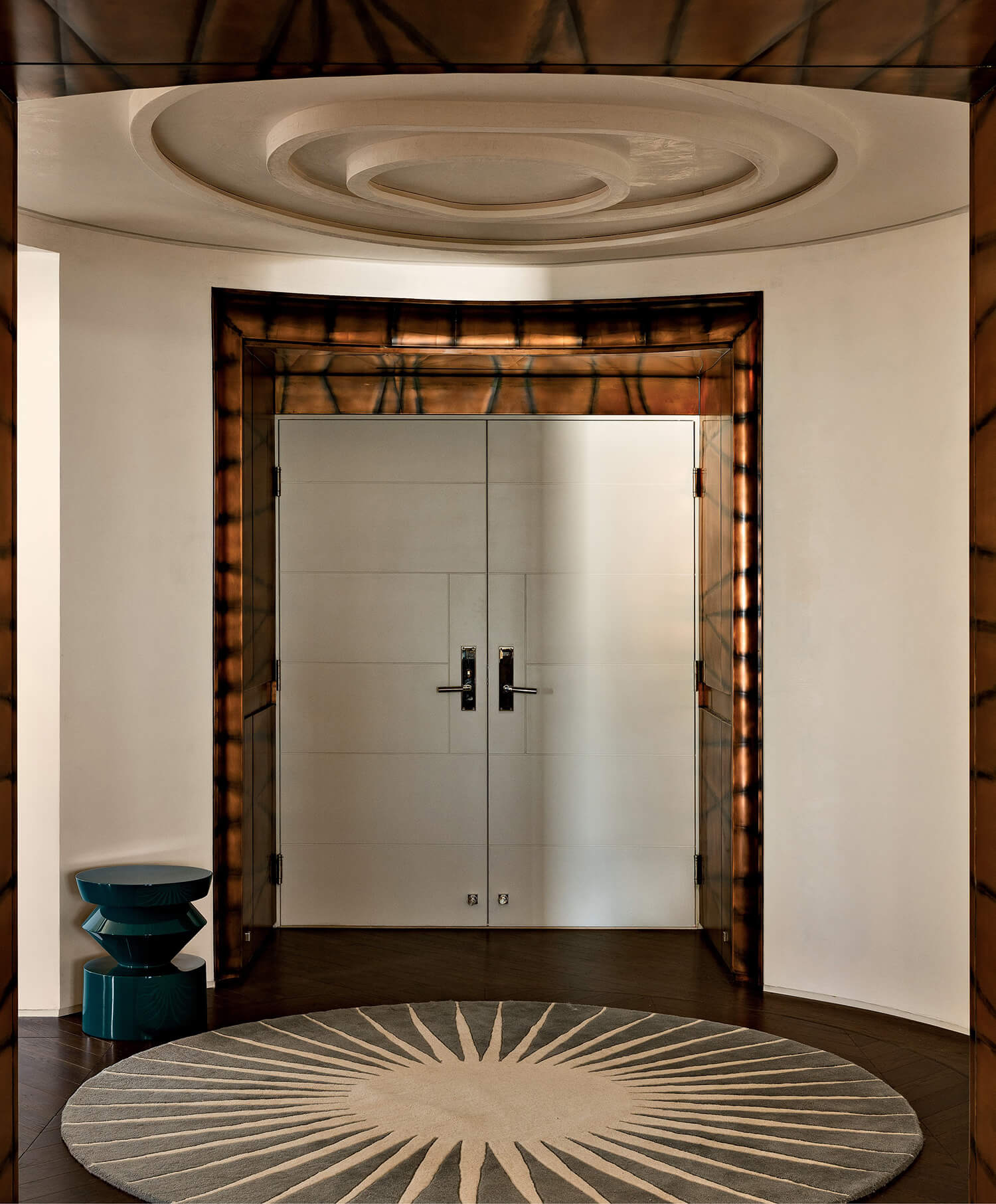 Double doors in a modern entryway with a round sunburst rug, geometric teal stool, and decorative ceiling and wall accents.