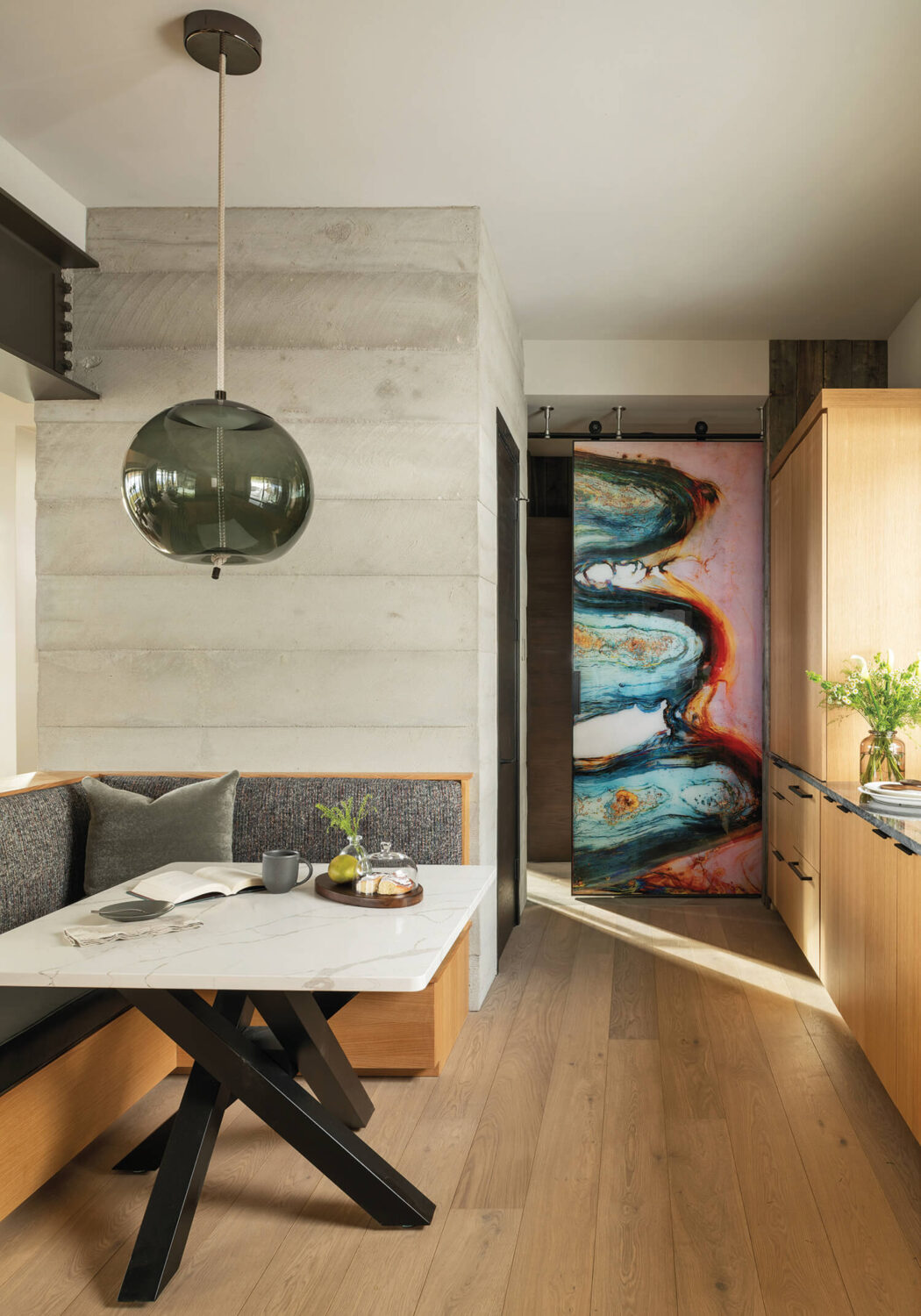 A modern kitchen with light wood floors, a marble-top table with bench seating, and a colorful abstract sliding door in the background.
