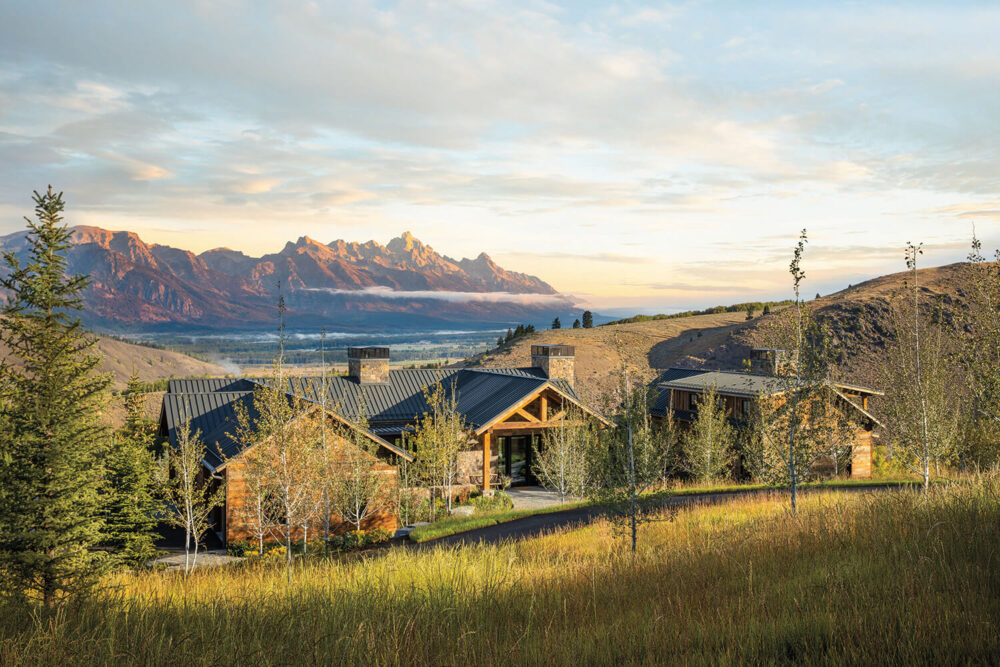 A rustic lodge-style house with a dark metal roof sits among trees and grass, with mountains and a partly cloudy sky in the background.