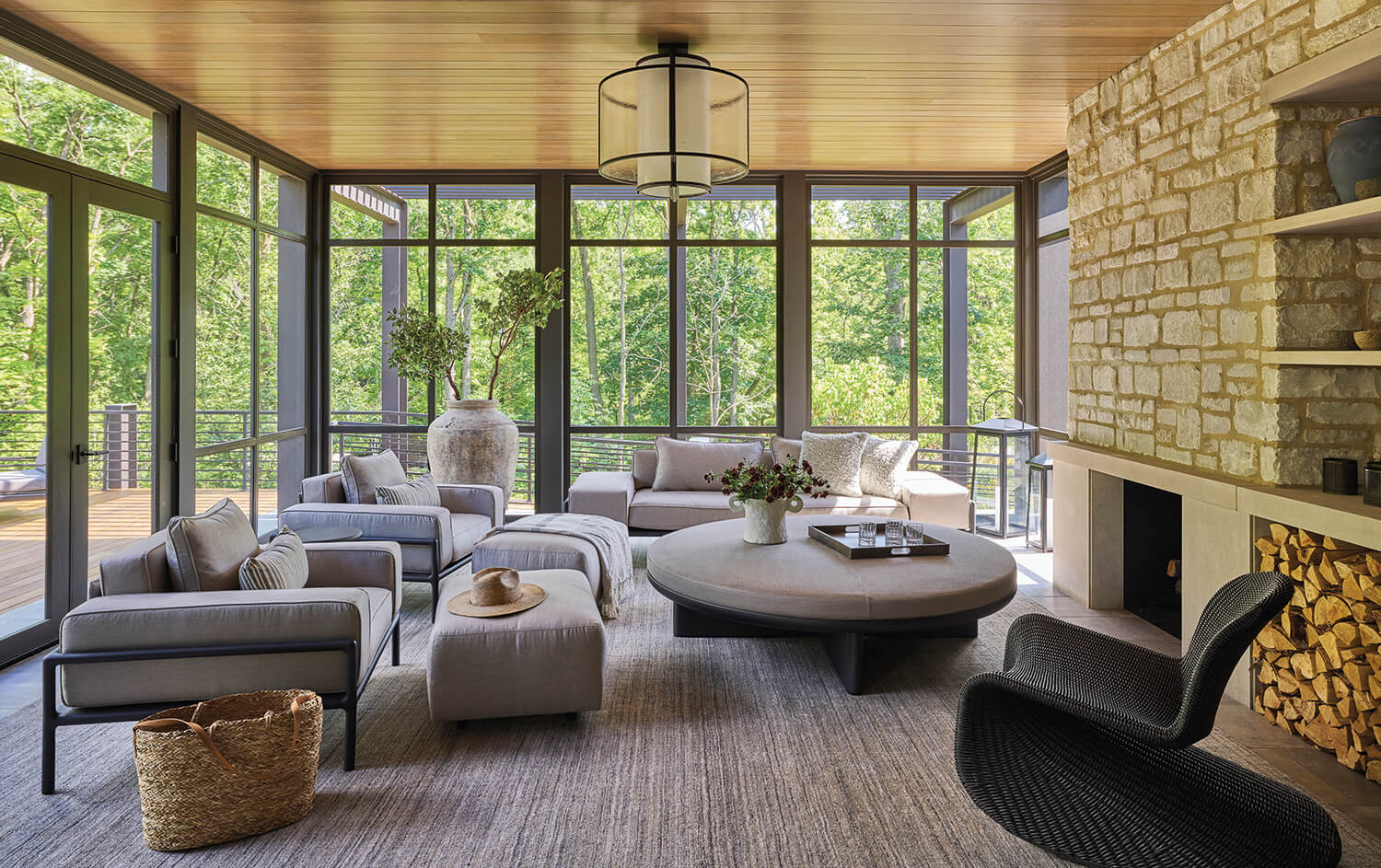 Modern living room with large windows, neutral-toned furniture, a round coffee table, a wicker basket, and a stone fireplace with stacked firewood. Trees are visible outside.