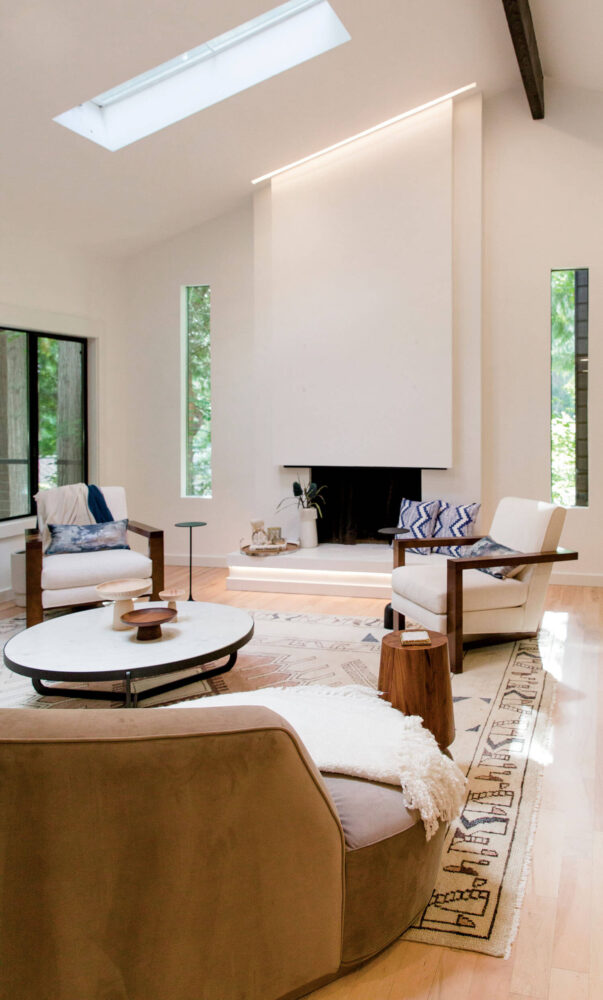 Modern living room with neutral armchairs, a round coffee table, patterned rug, and fireplace; large windows and skylight provide natural light.